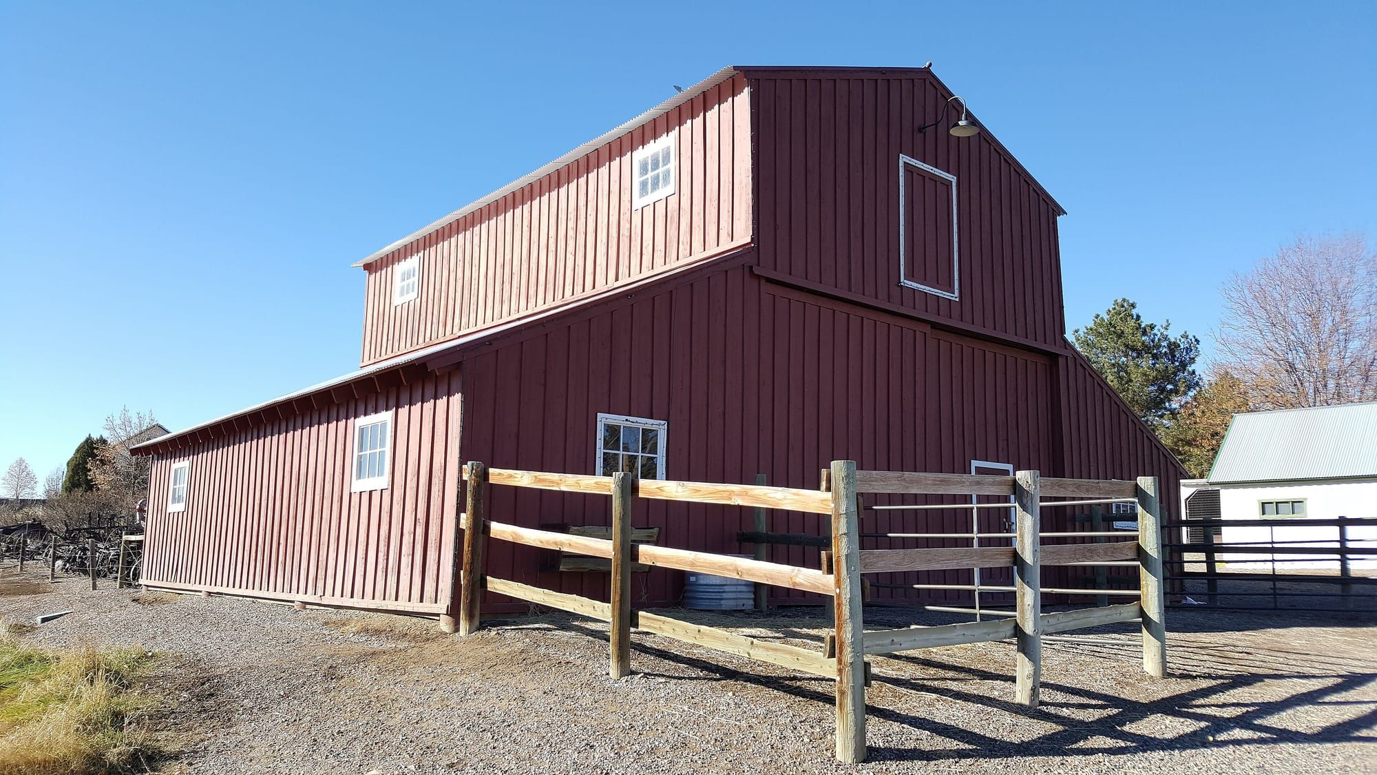 A large red-brown barn with a steep gambrel roof behind a wooden rail fence and a gravel yard, under a clear blue sky