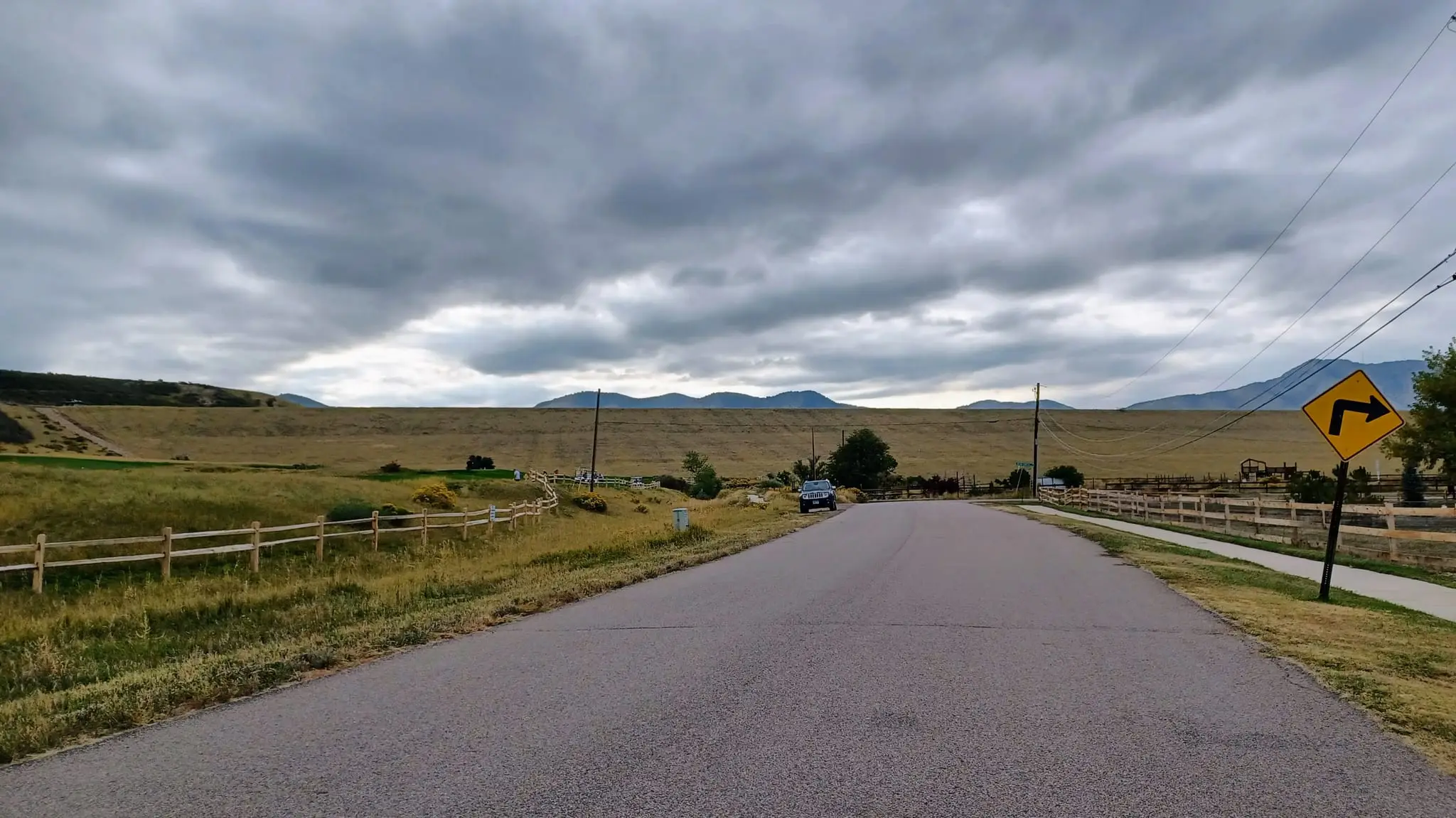 A paved path running beside a grass slope and a wooden fence toward distant hills, under a wide, heavy layer of grey cloud