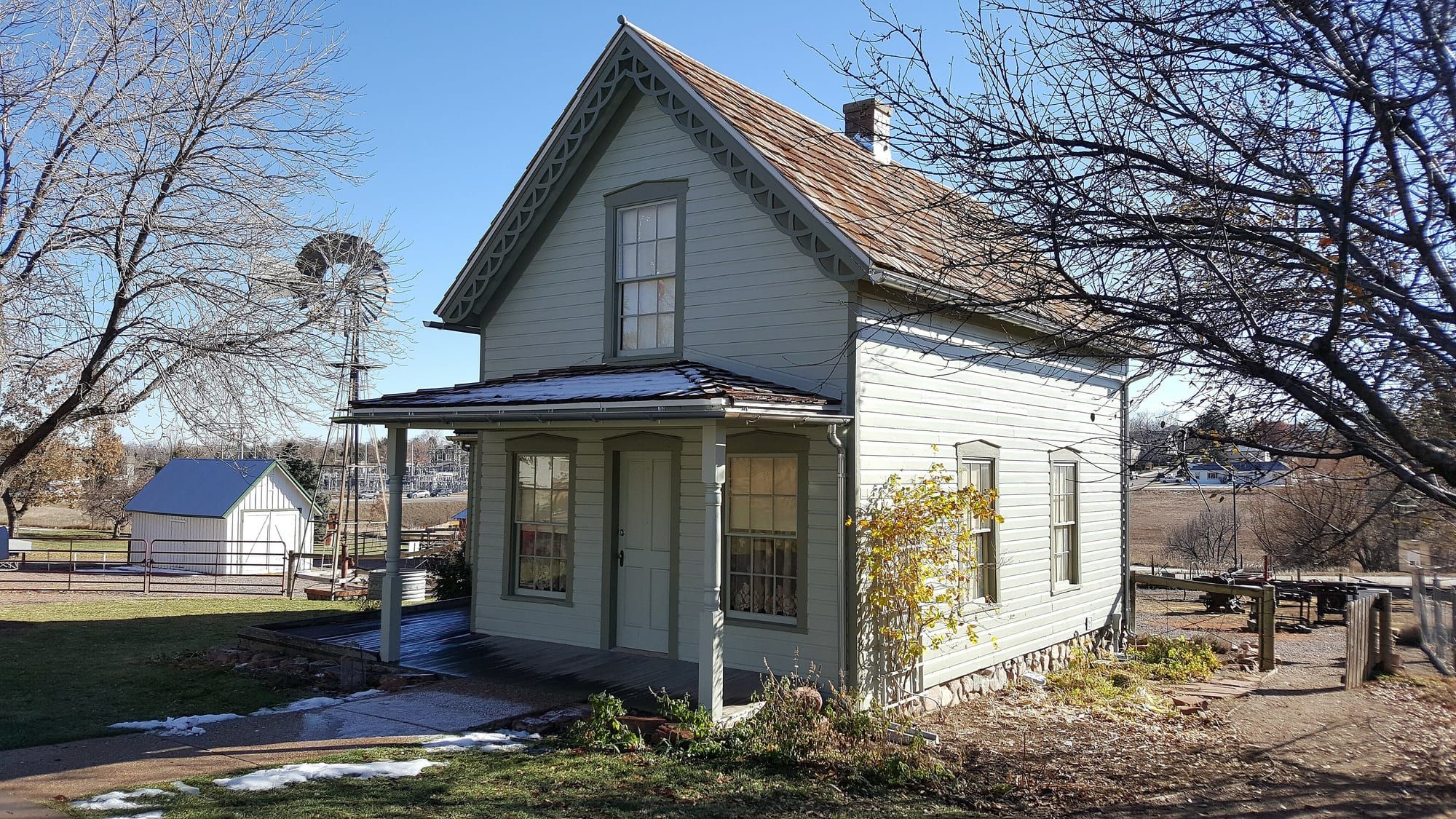 A pale wooden house with a shingled roof and a brick chimney, seen through bare trees in winter light